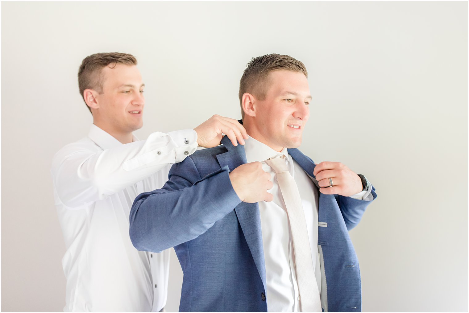 groomsman helps groom put on navy suit jacket for wedding