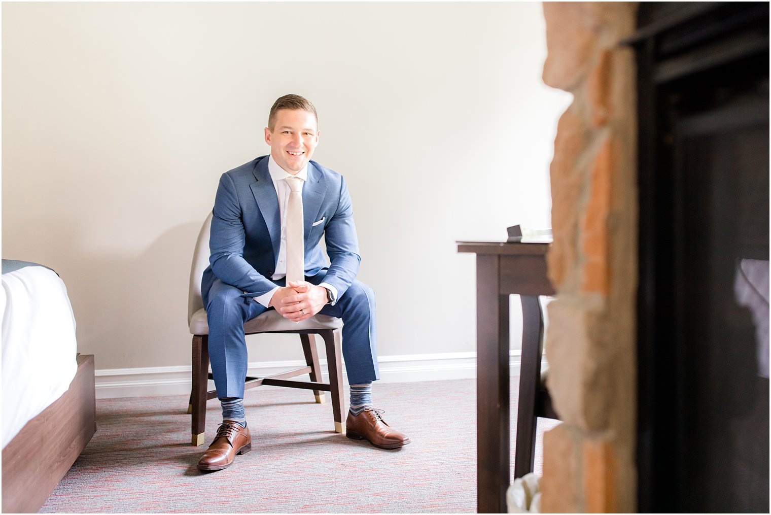 groom in navy suit sits on chair in NJ hotel