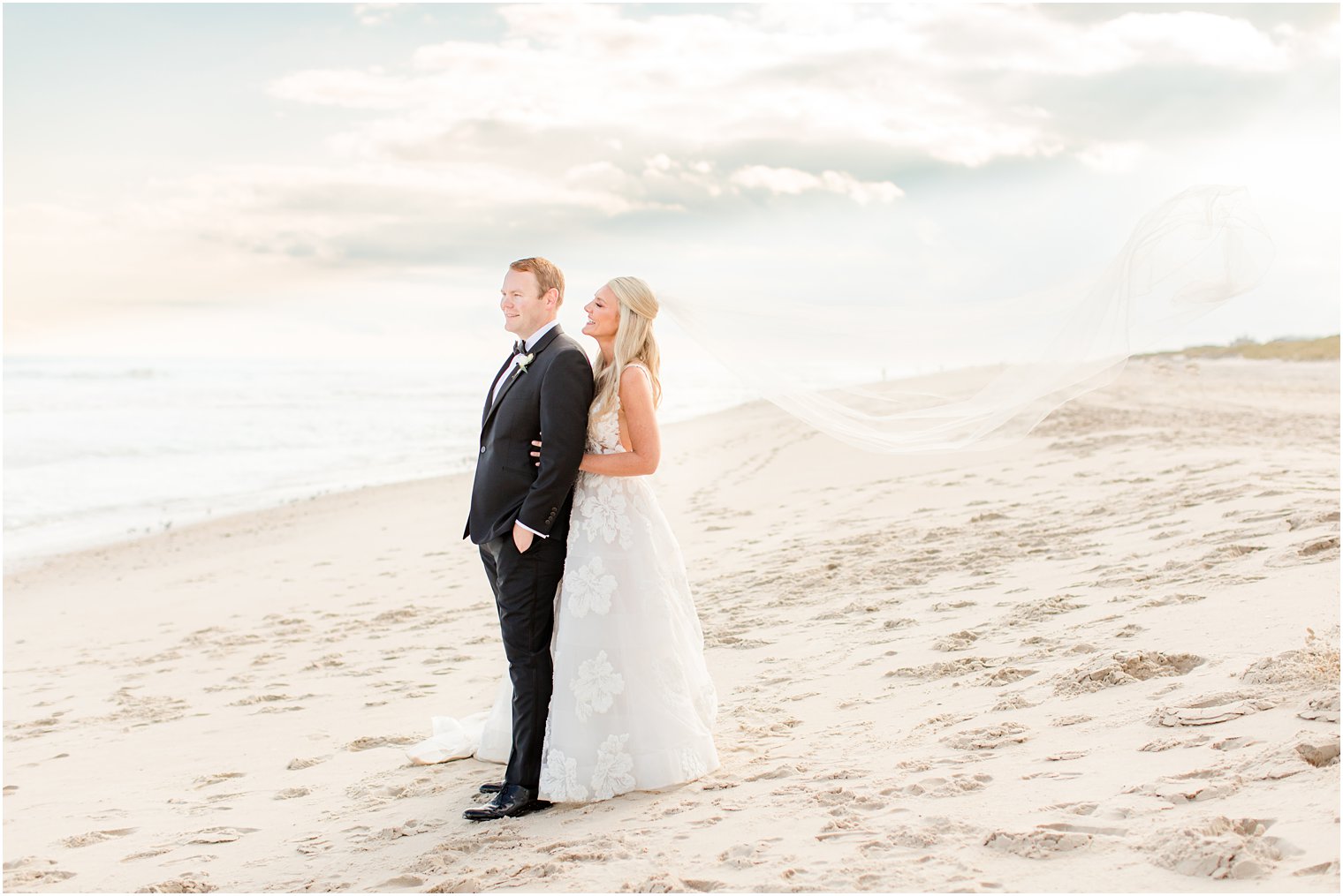 bride has groom from behind on beach with veil floating behind her