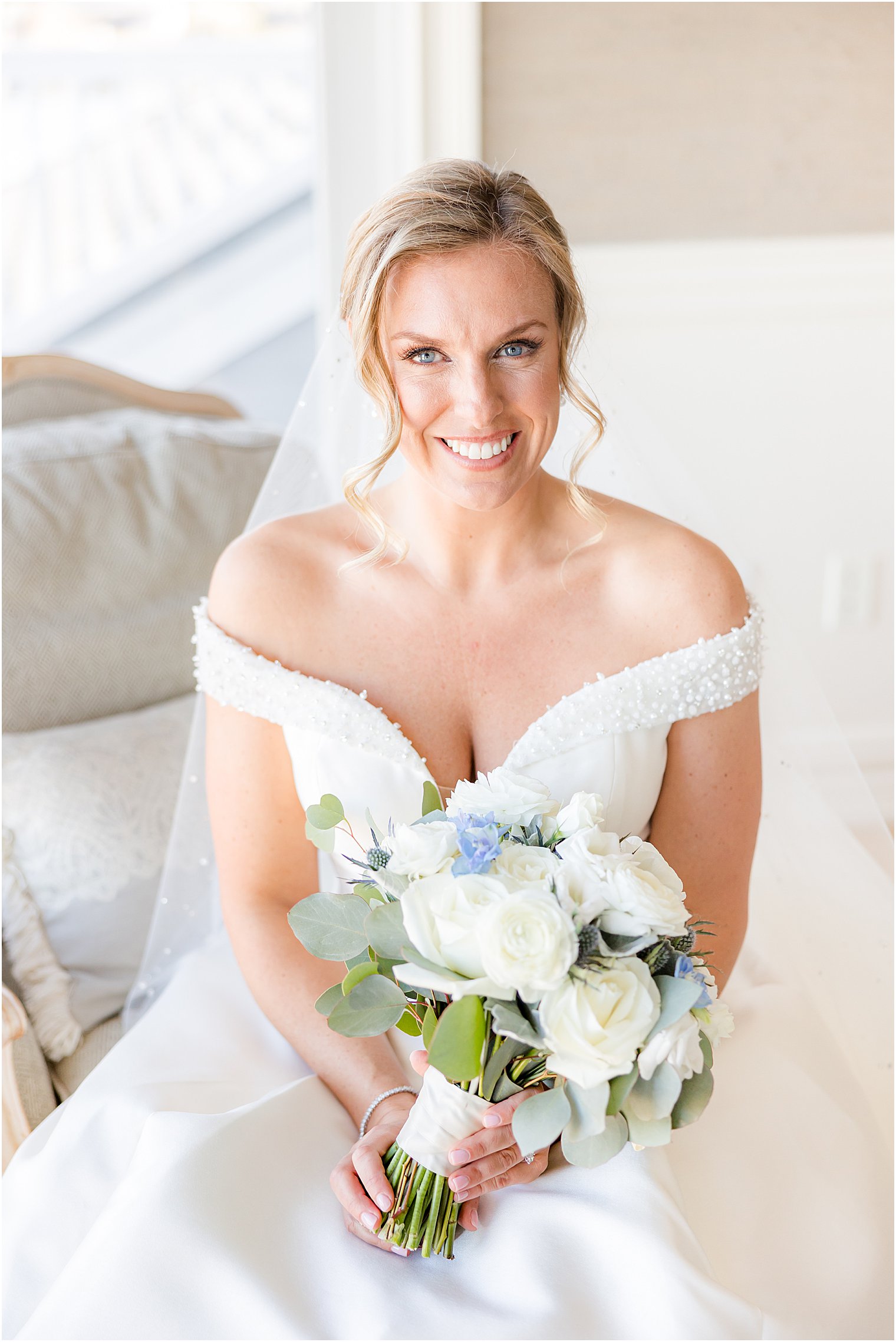 bride sits on chaise holding bouquet of white and blue flowers 